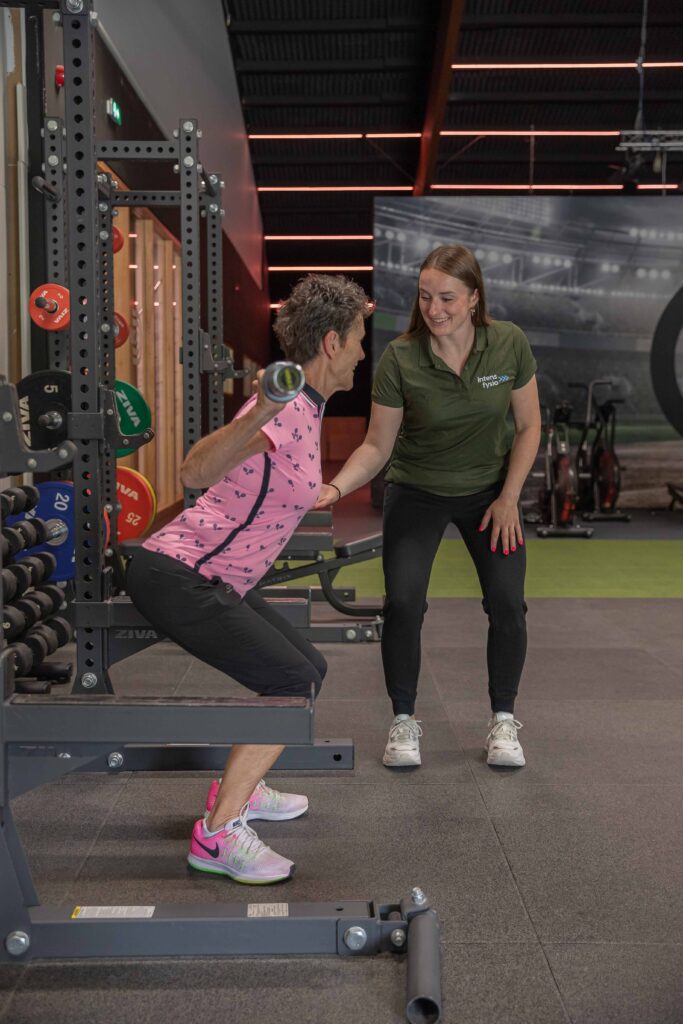 Fysiotherapeut ondersteunt senior tijdens squats met halter in de fitnessruimte van Intens Fit & Fysio